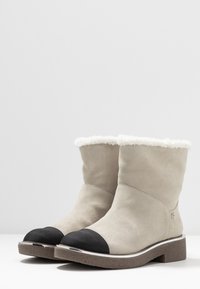 Botas de tobillo de ante en gris claro con puntera negra y forro de piel sintética blanca. Suela marrón texturizada con detalles en plata.