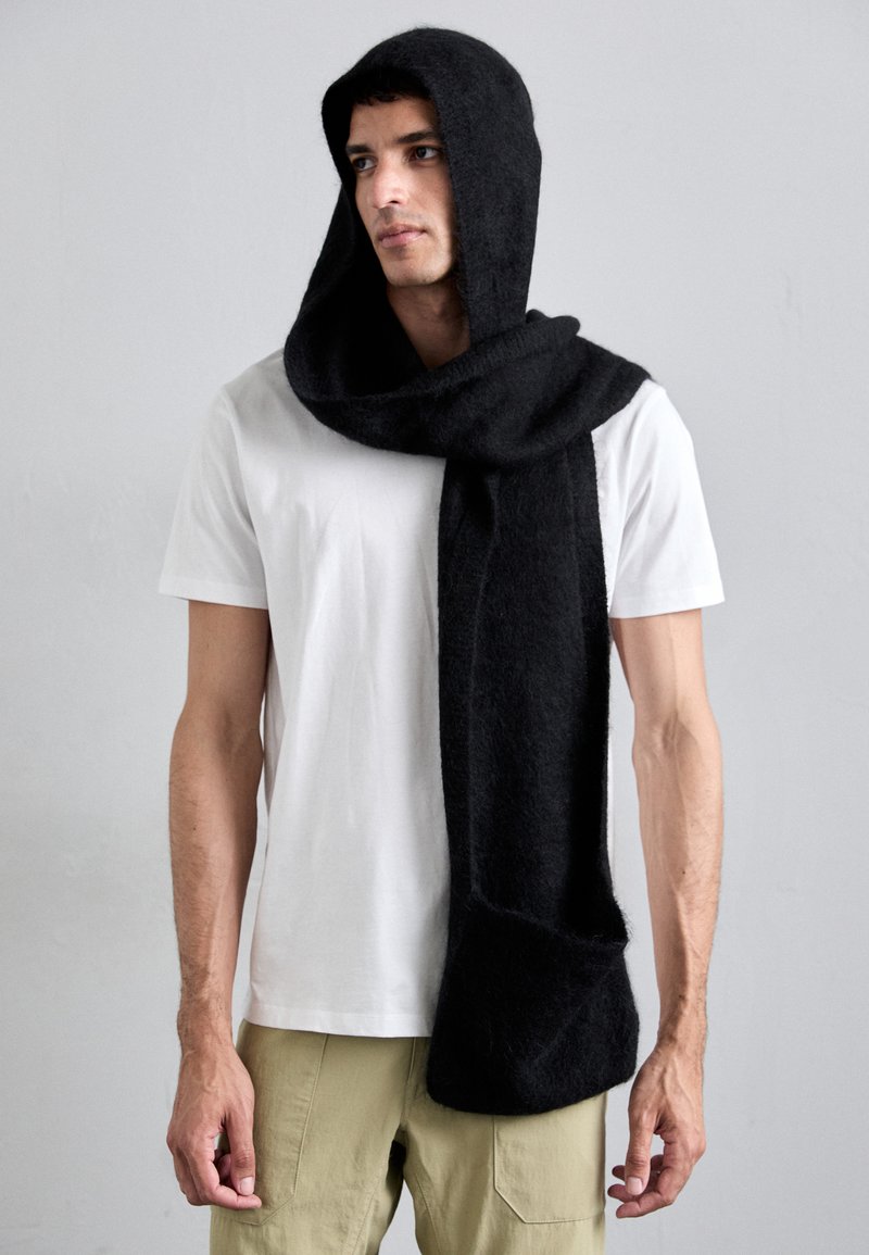 Écharpe-capuche en laine noire avec une finition douce et texturée, présentant un design drapé et une forme allongée. Portée par-dessus une chemise blanche.