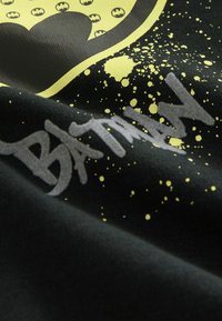 Čierne tričko z látky s žltým logom Batmana v štýle rozstreku a textom "BATMAN" v sivej farbe. Textúrovaný vzhľad a živé farby.