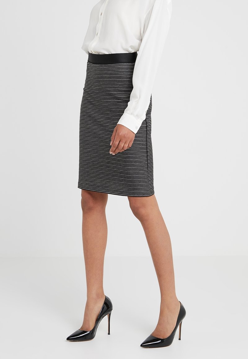 Femme portant une jupe crayon rayée noire et blanche, un chemisier blanc boutonné et des chaussures à talons noirs, debout sur un fond uni.