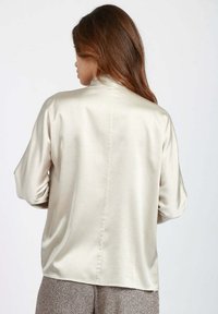Camicia in satin color beige chiaro con colletto alto, maniche lunghe e dettagli sulla cucitura posteriore. Texture liscia, vestibilità rilassata.