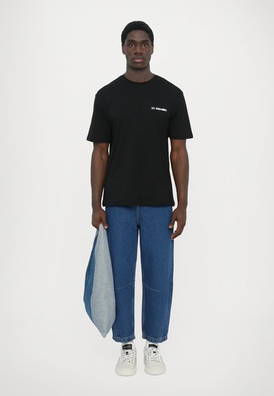 Μαύρο βαμβακερό κοντομάνικο μπλουζάκι με λευκό κείμενο στο στήθος, συνδυασμένο με μπλε φαρδιά τζιν παντελόνια και λευκά αθλητικά παπούτσια.