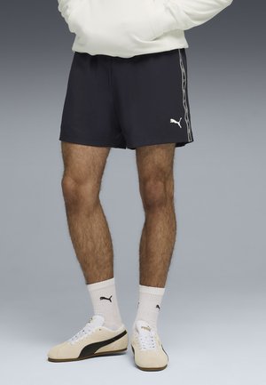 Mężczyzna w czarnych szortach Puma, białych skarpetkach Puma i beżowych butach Puma stoi na tle szarego tła.