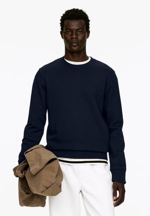 Dunkelblaues Sweatshirt mit rundem Halsausschnitt und langen Ärmeln, über einem schwarz-weißen gestreiften Hemd getragen. Beigefarbene Stoffjacke in der Hand gehalten.