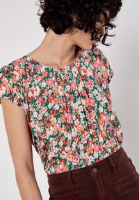 Blusa floreale a maniche corte, con uno sfondo verde e motivi floreali colorati in rosa, arancione e bianco. Dettaglio del collo arricciato.