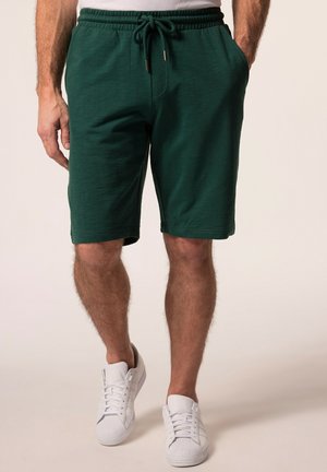 Mann trägt dunkelgrüne Shorts mit Kordelzug und weiße Sneakers, steht mit Händen in den Taschen vor hellem Hintergrund.