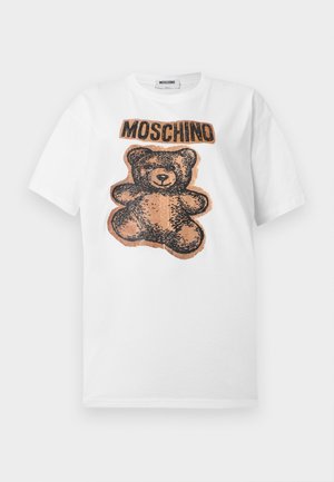 Vrouw draagt een wit t-shirt met een teddybeerdessin, lichtblauwe shorts met trekkoord, witte sokken en zwarte puntige hakken, staand met handen in de zakken.