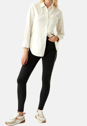 Kobieta w kremowej koszuli zapinanej na guziki, czarnych obcisłych dżinsach z bocznymi paskami i beżowych tenisówkach, z jedną ręką w kieszeni.