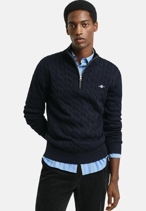 Pull en maille torsadée bleu marine avec un col montant et une fermeture éclair, superposé à une chemise à rayures bleues et un pantalon en velours côtelé noir.