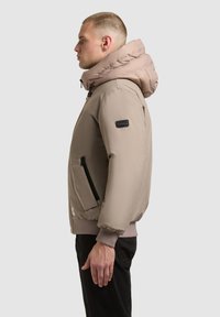 Világosbarna kapucnis, bézs bomberdzseki, cipzáras oldalzsebekkel és bordázott mandzsettareszel. Sima anyagú, karcsúsított fazon.