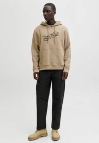 Mężczyzna w beżowym hoodie z grafiką, czarnych zwężających się spodniach i beżowych butach na sznurówki stoi na tle jasnym.