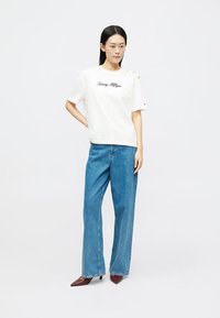 Hvit bomull T-skjorte med svart logo og knappdetaljer, parret med løse blå denimjeans og bordeaux spisse hæler.