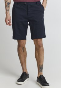 Shorts bleu marine avec un design à l'avant plat, deux poches latérales et une fermeture à bouton. Portés avec des baskets noires et des tatouages visibles sur les jambes.