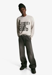 Svetlo šedý sveter s čiernym textovým dizajnom, uvoľnené tmavé denimové džínsy a čierne tenisky. Mäkko pletená čiapka dopĺňa outfit.