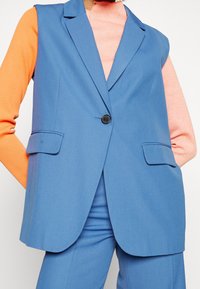 Blå ärmlös kavaj med skräddarsydd passform, en svart knapp, och två framfickor, tillsammans med en orange långärmad topp.