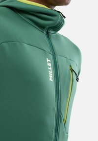 Green softshell jacket with a high collar, featuring a yellow accent on the neckline, zippered chest pocket, and the "Millet" logo on the front.