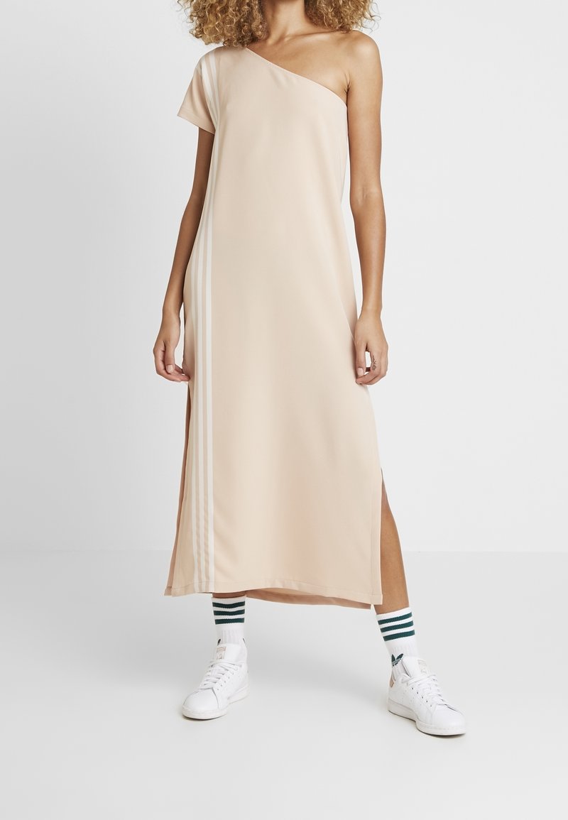 Beige jednopramenné šaty s diagonálním střihem, doplněné bočními rozparky, v kombinaci s bílými teniskami a zeleně pruhovanými pono žkami. Hladká textura látky.