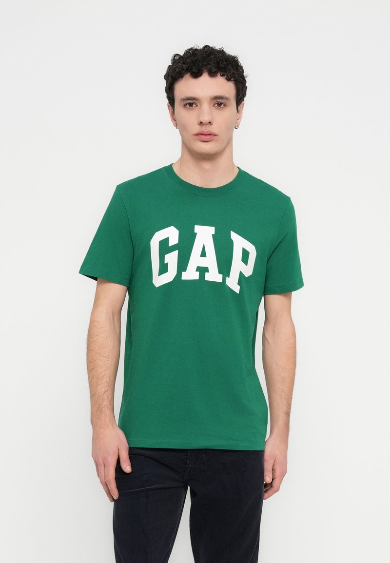 Πράσινο βαμβακερό t-shirt με μεγάλο λευκό λογότυπο "GAP" στο μπροστινό μέρος. Μανίκια κοντά και στρογγυλή λαιμόκοψη, παρουσιάζεται σε απλό φόντο.