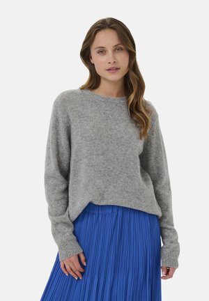 Junge Frau mit langen, welligen Haaren, die einen hellgrauen Pullover und einen leuchtend blauen Plissée-Rock trägt, steht vor einem schlichten Hintergrund.