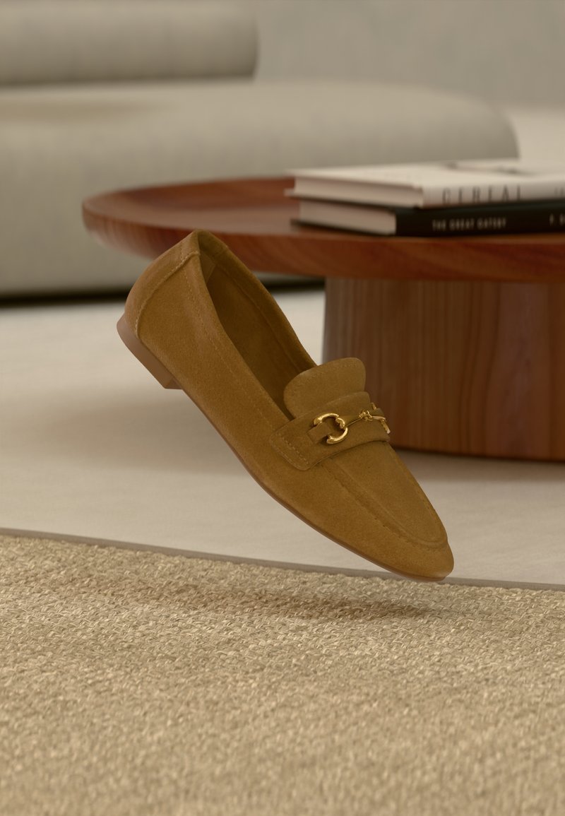 Mocassin en daim marron avec détail mors doré flottant au-dessus d'un tapis beige texturé, avec une table basse en bois et des livres en arrière-plan.