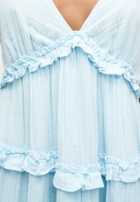 Ανοιχτό μπλε ύφασμα με κάθετες ρίγες που διαθέτει διακοσμητικές βολάν κατά μήκος του V-λαιμού και της μέσης σε ένα ένδυμα.