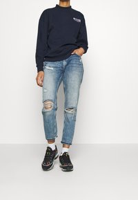 Marinblå sweatshirt med hög krage, ljusblå slitna jeans med hål, och svarta sneakers med färgglada detaljer.