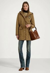 Donna che indossa una giacca imbottita beige, un maglione a fantasia, jeans blu e stivali ankle brown, mentre tiene in mano una borsa in pelle marrone su uno sfondo bianco.