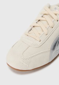 Scarpa in suede bianca con una striscia laterale argento, dotata di lacci piatti e suola testurizzata. Dettaglio notevole: logo impresso nella zona delle punte.