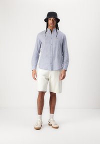 Blå og hvid stribet langærmet skjorte med knapper, cremefarvede shorts, hvide sneakers med brune såler og en sort bøttehat. Lige design.