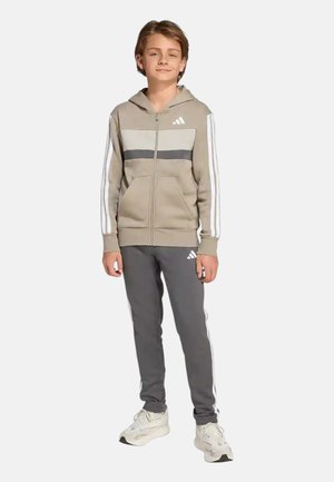 Ragazzo in piedi che indossa una felpa con cappuccio con cerniera beige e grigia Adidas e pantaloni della tuta grigi Adidas con sneaker bianche, su sfondo semplice.