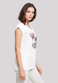 Weißes Baumwoll-T-Shirt mit einem grafischen Druck eines Skeletts, das herzförmige Luftballons hält. Kurze Ärmel, entspannter Schnitt, glatte Textur.