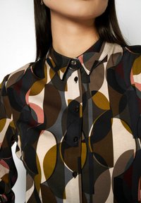 Bluse mit Knopfverschluss und geometrischem Muster in Erdtönen, mit spitzem Kragen und schwarzen Knöpfen auf der Vorderseite. Glatte Stofftextur.