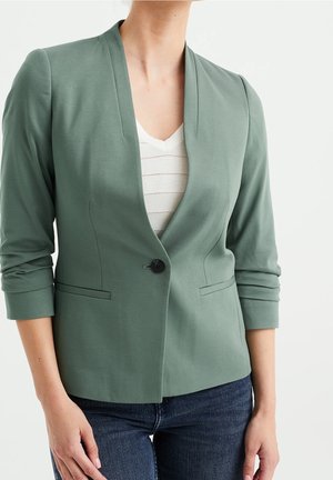 Frau trägt salbeigrünen Blazer mit einem schwarzen Knopf, weißes V-Ausschnitt-Top und blaue Jeans, Hände locker an den Seiten, neutraler Hintergrund.