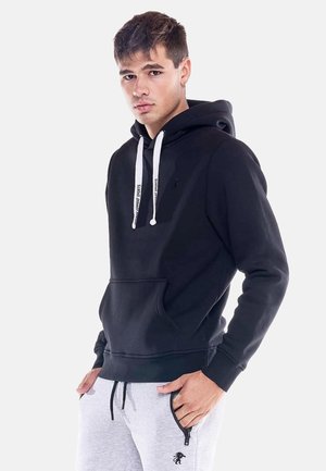 Jeune homme aux cheveux courts et foncés portant un sweat à capuche noir avec des cordons blancs et un pantalon de survêtement gris clair avec une poche zippée noire.
