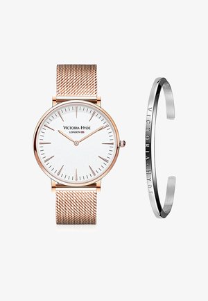 Montre en maille dorée rose avec un cadran blanc, des marqueurs d'heure minimalistes et un bracelet noir avec des lettres en relief. Les deux articles présentent un design épuré.