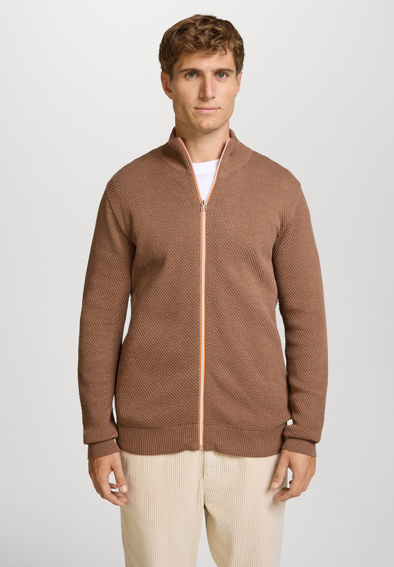 Brun lynlåsjakke med et tekstureret mønster, kontrasterende orange lynlås, ribbede manchetter og kant, båret over en hvid skjorte.