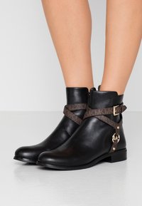 Bottines en cuir noir avec une sangle texturée marron et un détail de boucle dorée. Semelle plate et design à bout arrondi ; accent logo sur le côté.