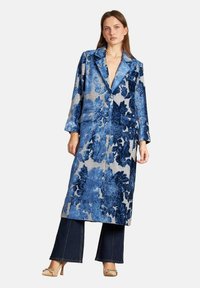 Abrigo largo azul con estampado floral y textura brillante, que presenta un cuello de solapa, botones de doble fila y dos bolsillos frontales, combinado con vaqueros oscuros.