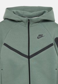 Sweatshirt verde da Nike com fecho na frente, capuz, riscas em forma de chevron pretas e pequeno bolso com fecho na manga esquerda.