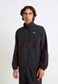 Svart zip-up jakke med røde detaljer og Puma-logo. Har høy krage og elastiske mansjetter, laget av lett, teksturert stoff.