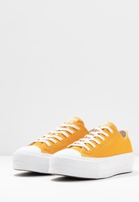 Paire de baskets basses en toile jaune avec semelles en caoutchouc blanches et lacets blancs sur une surface blanche.
