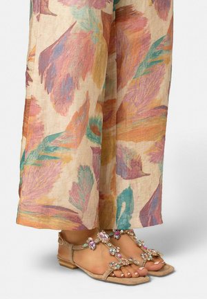 Pies con sandalias beige adornadas con piedras preciosas multicolores, combinadas con pantalones de pierna ancha con un estampado abstracto de hojas en tonos pastel.