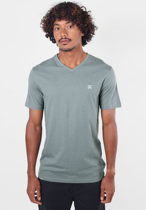 Homme aux cheveux bouclés portant un t-shirt vert clair à col en V et un pantalon noir, debout et face à l'avant sur un fond uni.