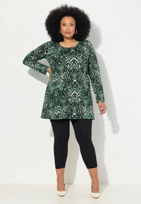 Tunique verte à motifs avec manches longues, présentant un motif tourbillonnant. Associée à des legging noirs et des talons beiges, sur un fond neutre.