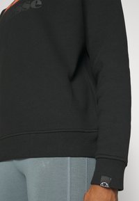 Gros plan d'une personne portant un sweat-shirt noir à manches longues et un pantalon bleu-gris, montrant les détails des poignets et des coutures latérales.
