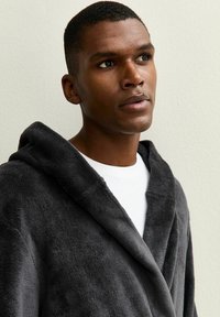 Charcoalgrijze badjas met capuchon in zachte, pluizige stof met een sjawlboord. Het model draagt een wit shirt eronder. Gladde textuur, ontspannen pasvorm.