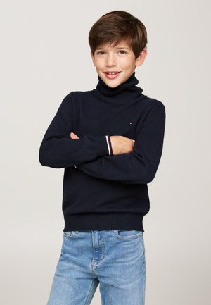 Maglione a collo alto blu navy con polsini e orlo a coste, caratterizzato da un piccolo logo. Indossato con jeans in denim azzurro chiaro.