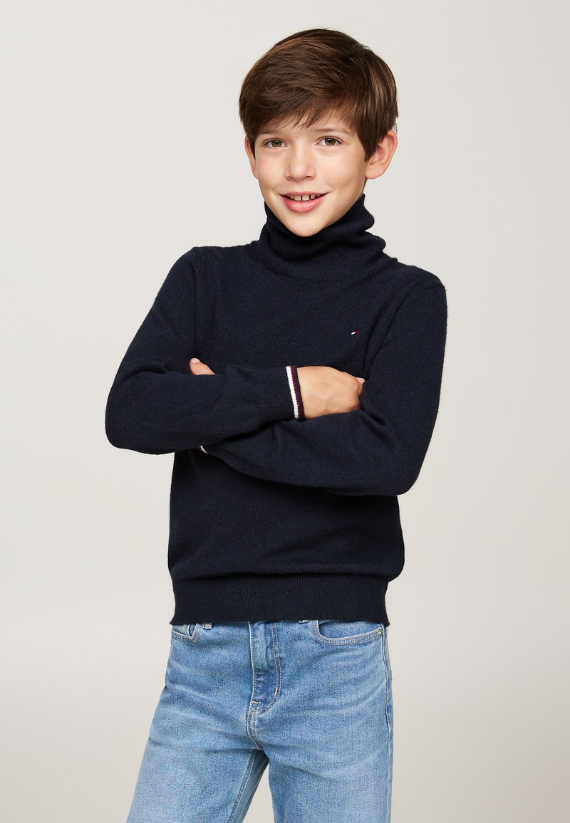 Maglione a collo alto blu navy con polsini e orlo a coste, caratterizzato da un piccolo logo. Indossato con jeans in denim azzurro chiaro.