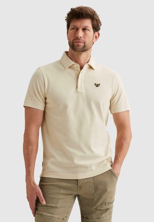 Mann trägt ein beiges Poloshirt mit kurzen Ärmeln und kleinem Flügellogo sowie olivfarbene Cargo-Hosen, steht mit einer Hand in der Tasche.