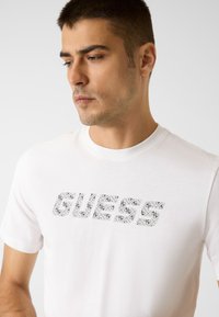 Weißes Baumwoll-T-Shirt mit kurzen Ärmeln und großem "GUESS"-Logo in schwarz-weißen Mustern auf der Brust. Glatte Textur.